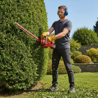A person safely operating a hedge trimmer with a rotating handle, wearing full PPE (safety glasses, gloves, ear protection), trimming a hedge with proper posture